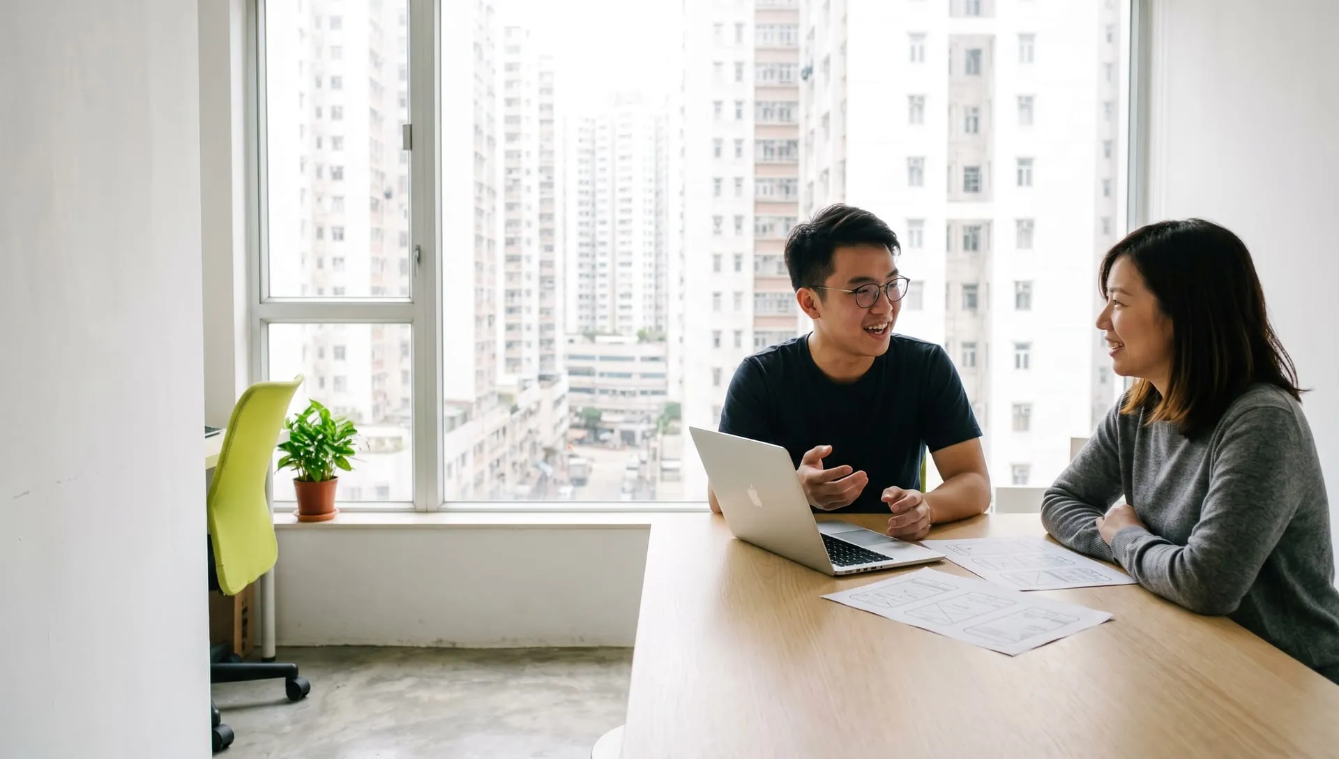 HIMX 開發者在明亮工作室與客戶隔著工作枱面對面傾談,桌上放著手提電腦和線框圖,窗外是香港樓宇